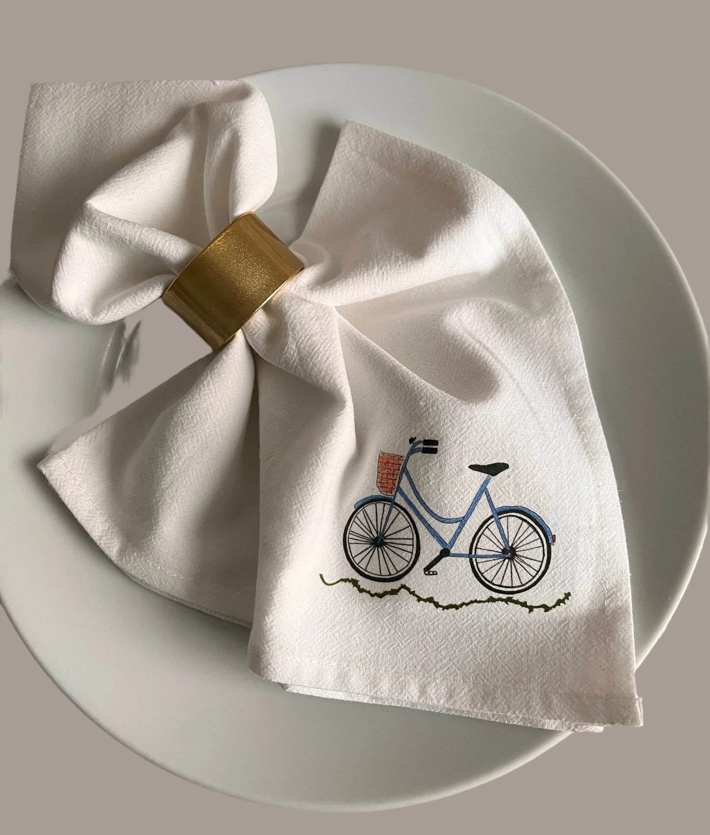 Cotton napkin with blue bicycle illustration and gold napkin ring on white plate