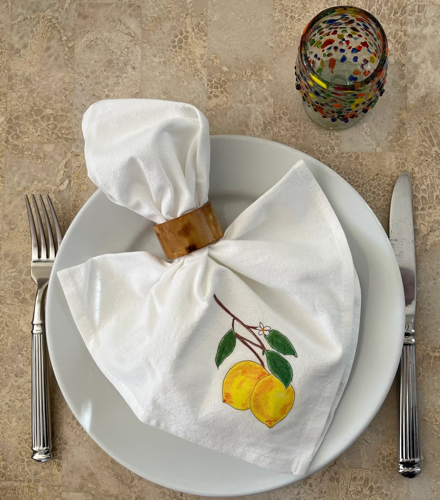 Illustrated lemon cotton napkin with napkin ring on plate, fork, knife, and colorful glass