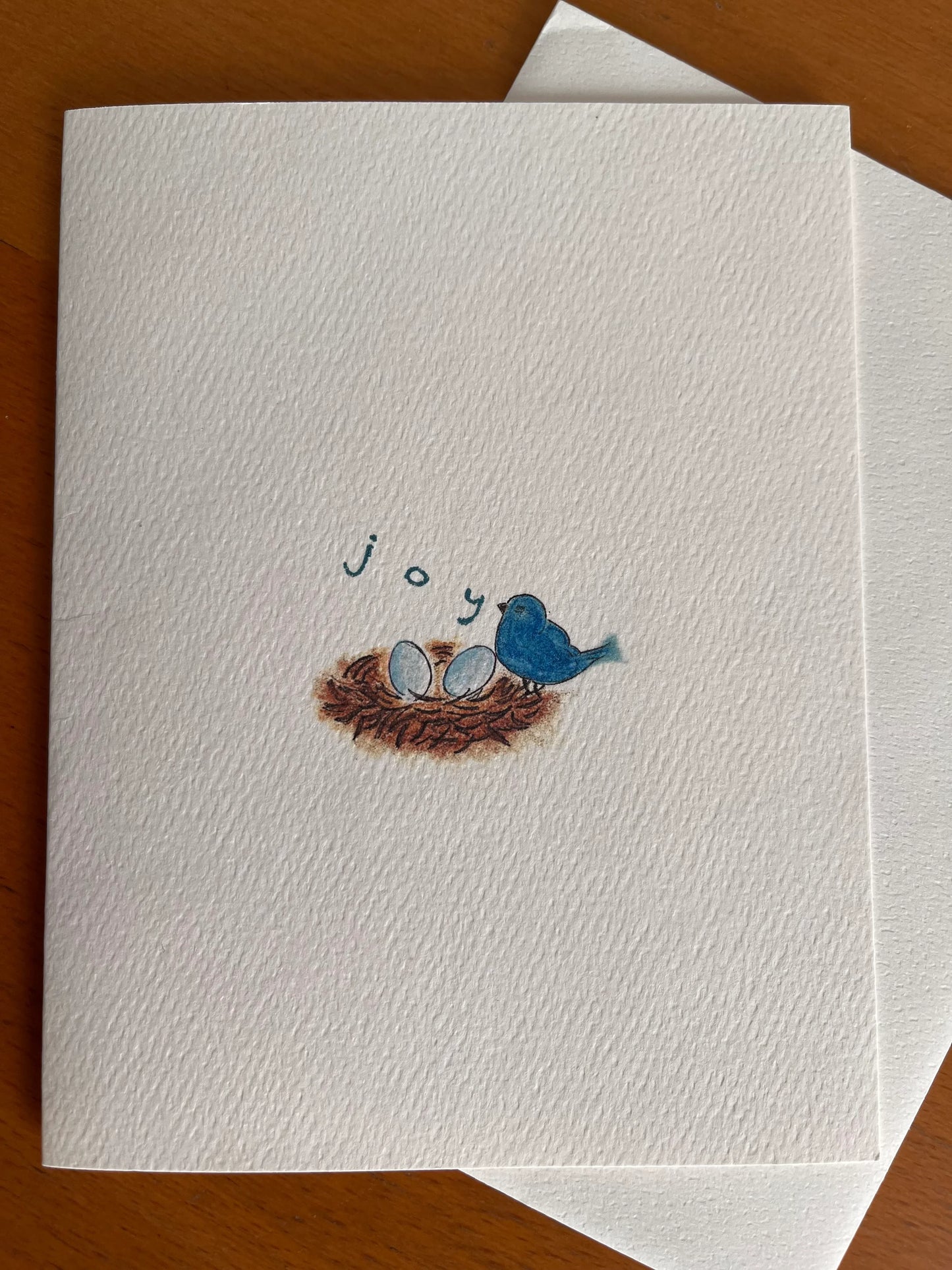 Illustrated note card with bluebird, nest, eggs, and 'joy' on textured paper