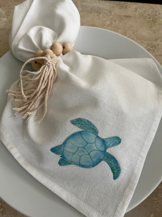 White cotton napkin with illustrated blue sea turtle, tied with wooden bead and tassel ring on plate