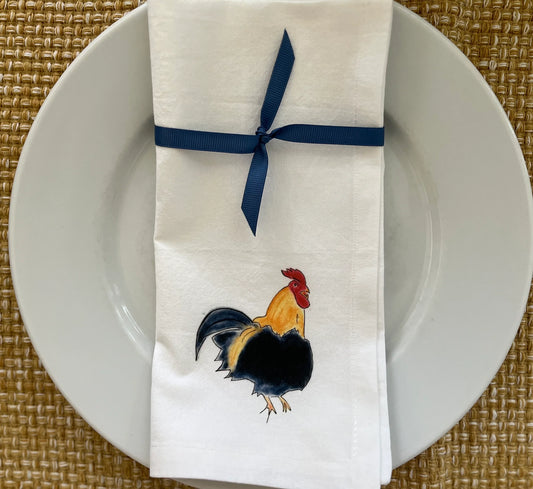 Cotton napkin with rooster illustration, tied with blue ribbon on white plate, woven placemat