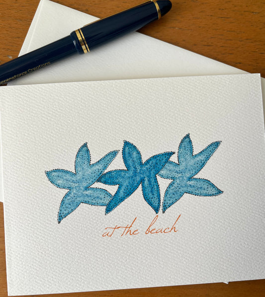 Note card with blue watercolor starfish illustration and 'at the beach' text, pen on table