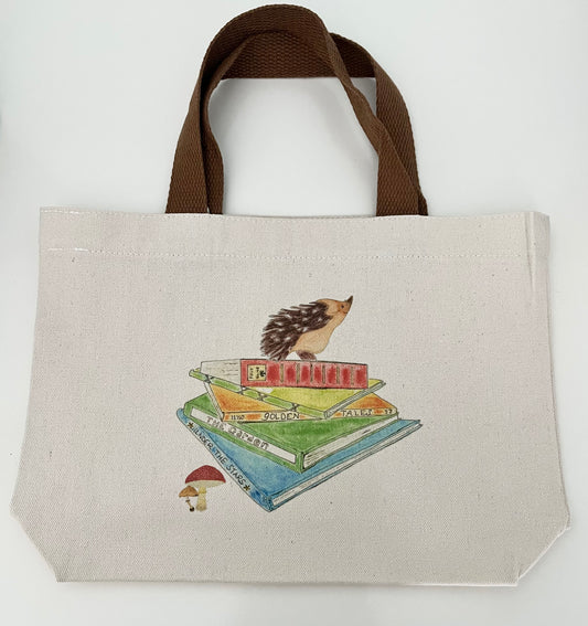 Canvas tote bag with brown handles, featuring a hedgehog illustration atop stacked books