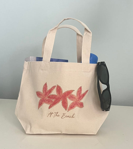 Beach tote bag with pink starfish print and 'At The Beach' text, holding sunglasses and a water bottle