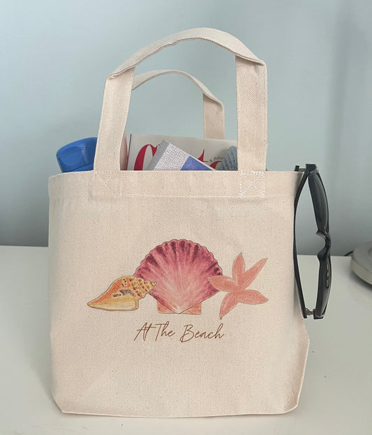 Beach-themed canvas tote bag with seashell, starfish, and 'At The Beach' text, perfect for seaside outings.