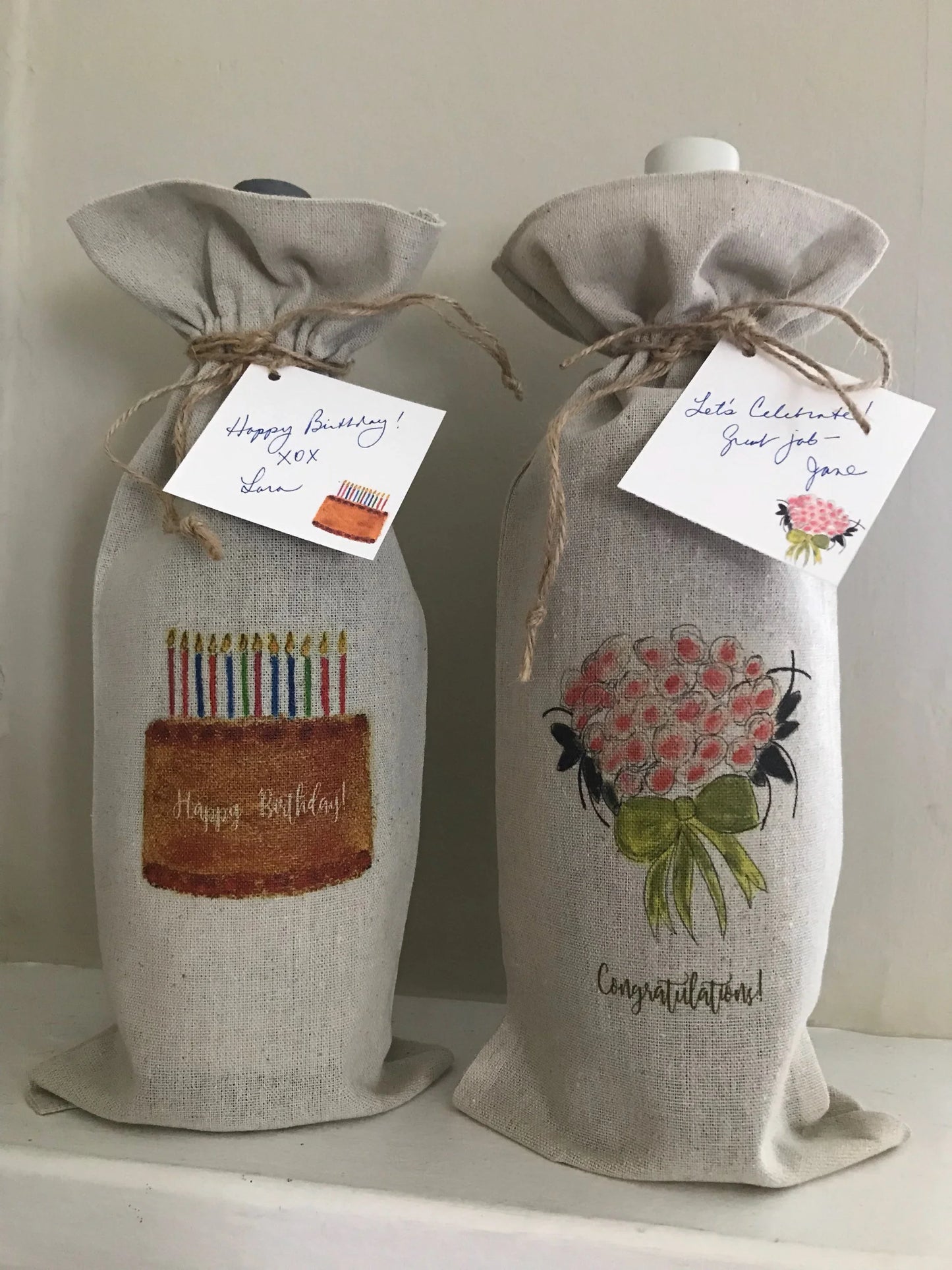 Two illustrated linen wine gift bags with birthday cake and bouquet designs, tied with twine.