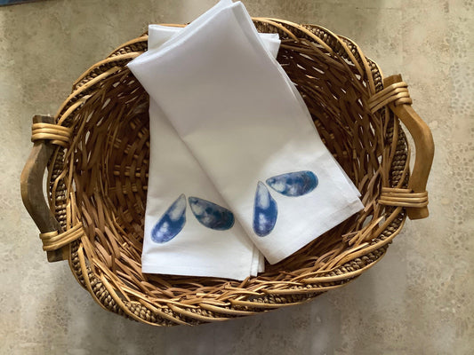 White cotton tea towels with blue watercolor shell illustrations in a wicker basket