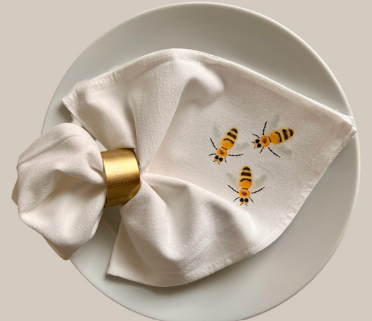 Cotton napkin with illustrated bees, gold napkin ring, displayed on a white plate