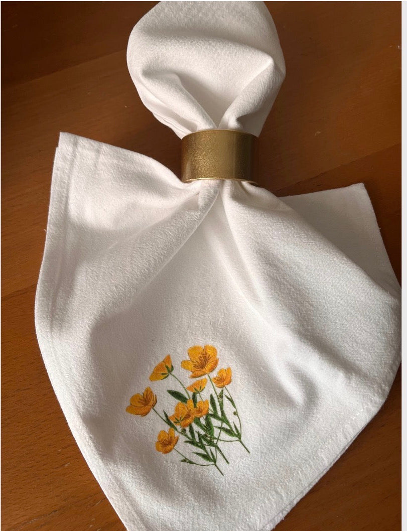 White cotton napkin with yellow wildflower illustration and gold napkin ring on wooden table