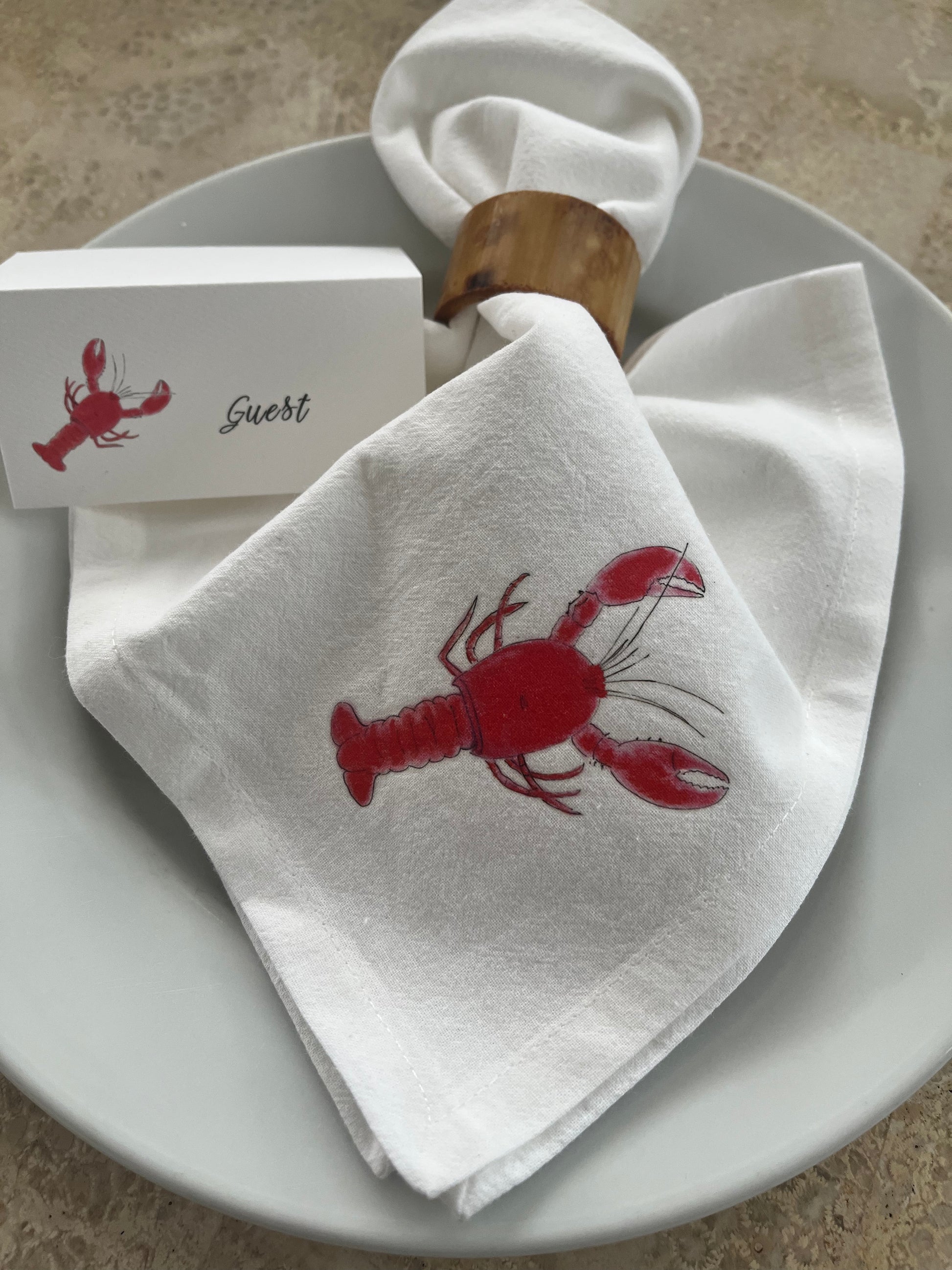 White cotton napkin with red lobster illustration, bamboo napkin ring, and guest place card on plate