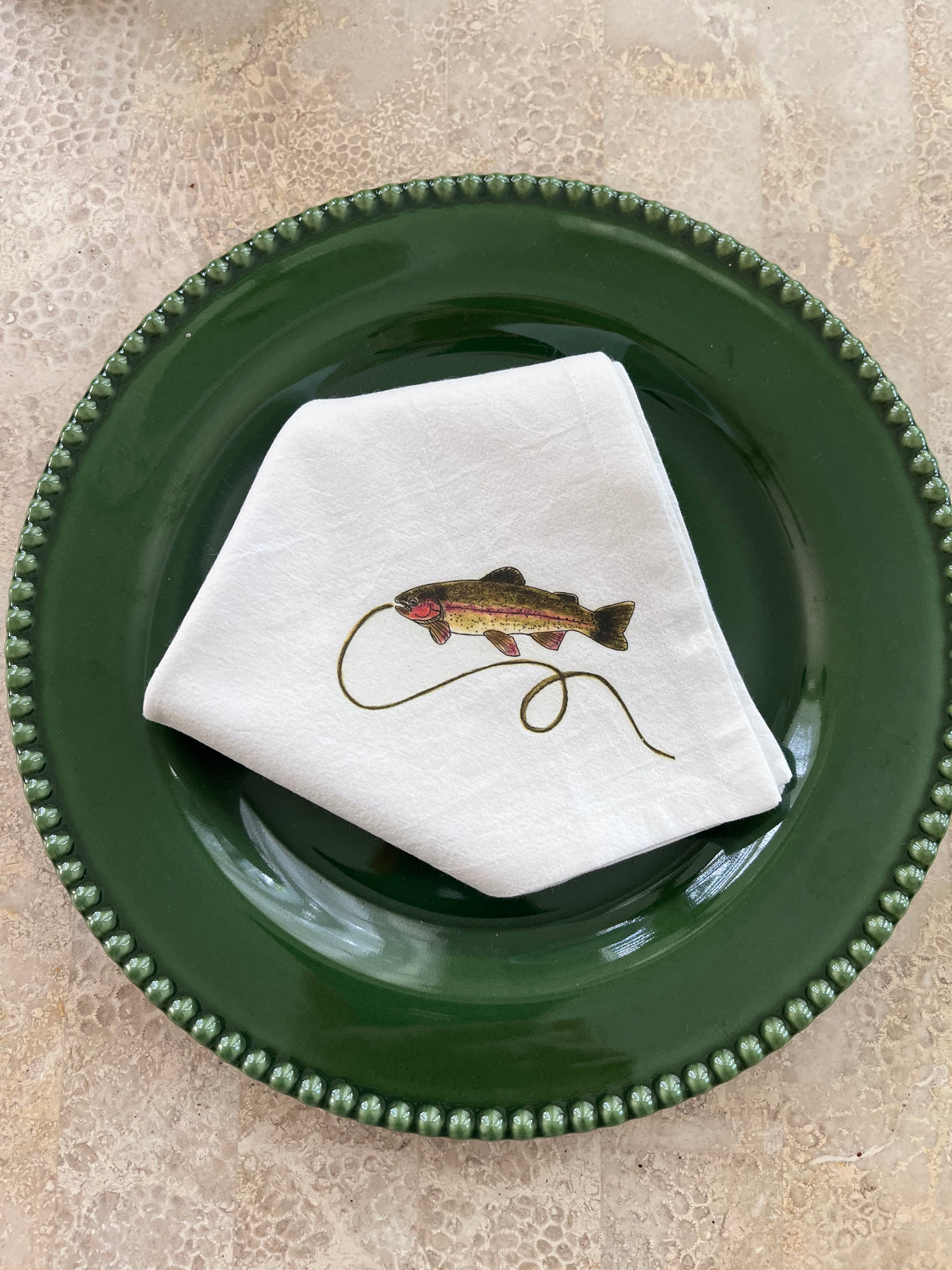 White cotton napkin with illustrated rainbow trout and fishing line, styled on a green beaded plate for nature-themed table settings.
