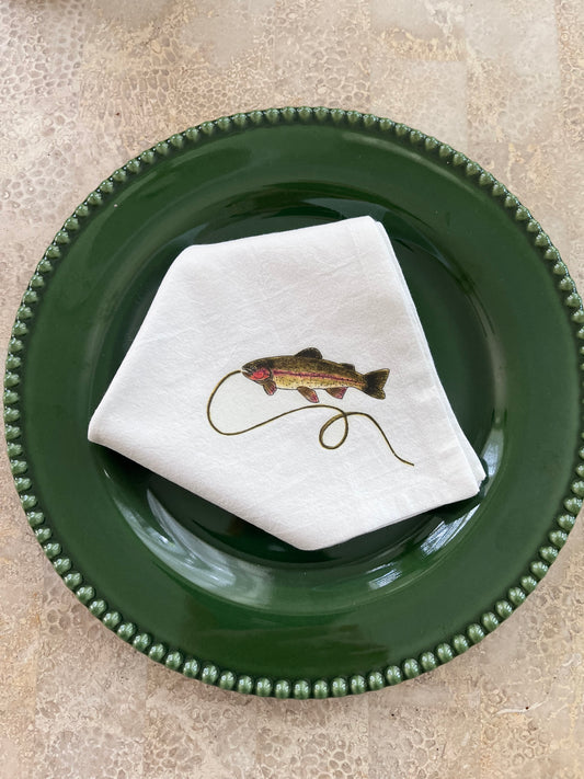 White cotton napkin with illustrated rainbow trout and fishing line, styled on a green beaded plate for nature-themed table settings.