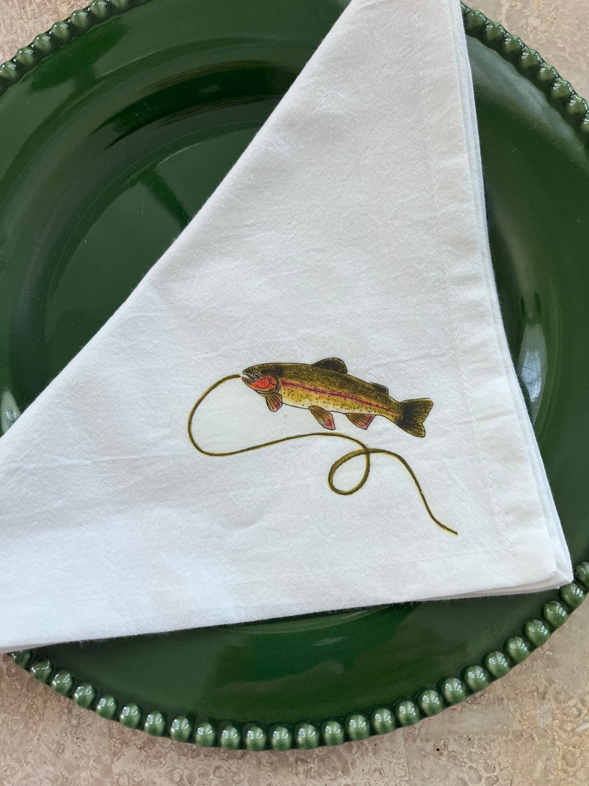 White cotton napkin with illustrated rainbow trout and fishing line, styled on green beaded plate for nature-inspired table settings.