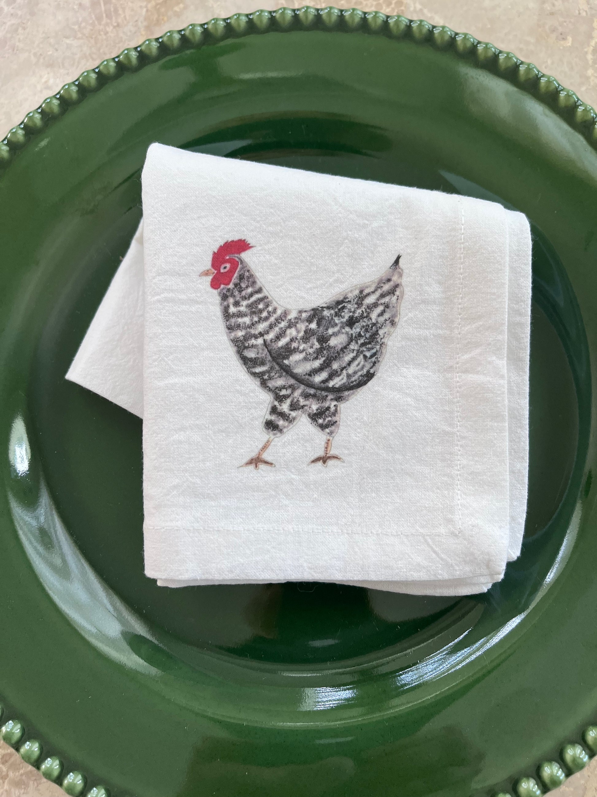 White cotton napkin with illustrated rooster, placed on green beaded plate for farmhouse table setting