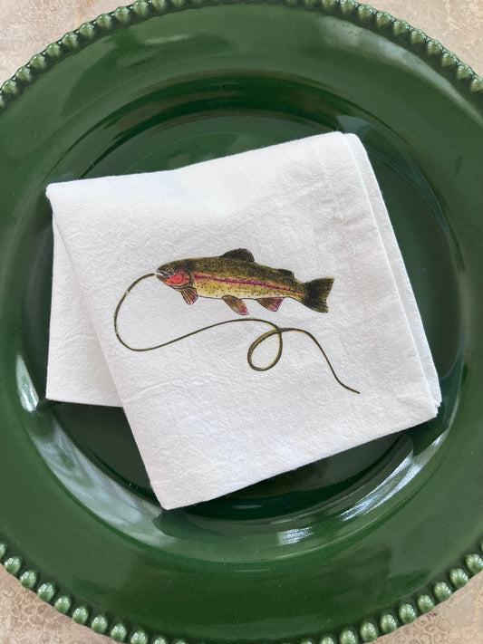 White cotton napkin with illustrated rainbow trout and fishing line, styled on green beaded plate for nature-inspired table settings.