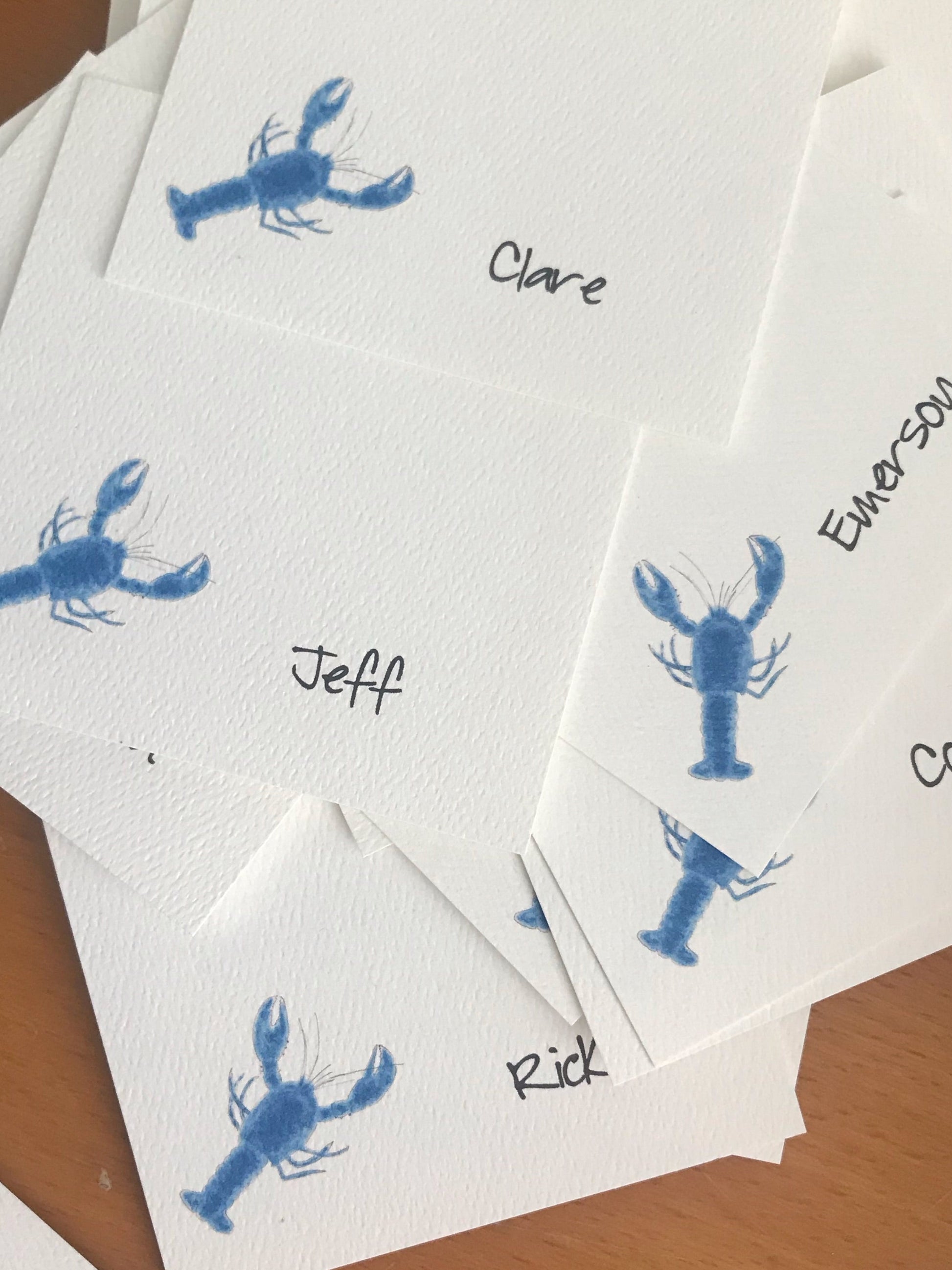 A stack of white textured paper place cards printed with a blue lobster illustration and handwritten names, arranged on a wooden surface.