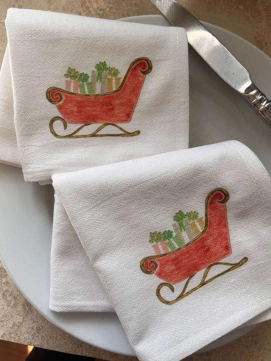 White cotton napkins with red sleigh design carrying potted plants, set on a plate with knife for festive table settings.