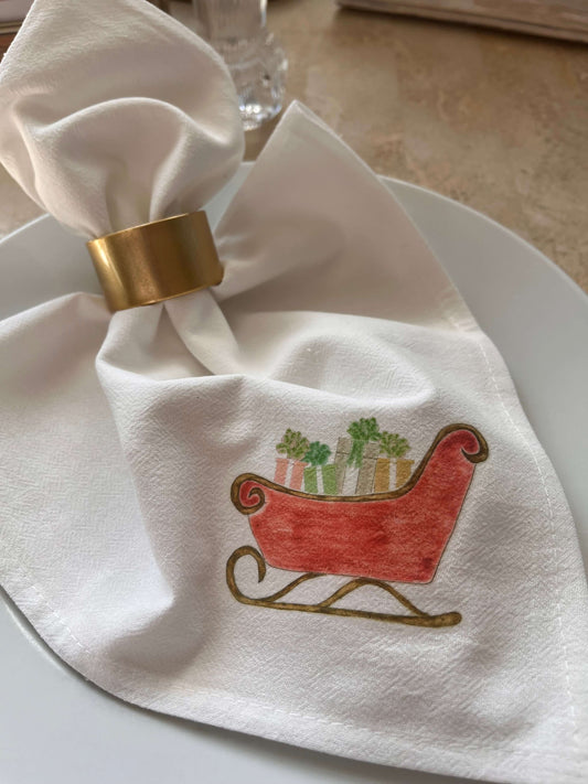 White cotton napkin with red sleigh & gift illustration, gold napkin ring, holiday table setting
