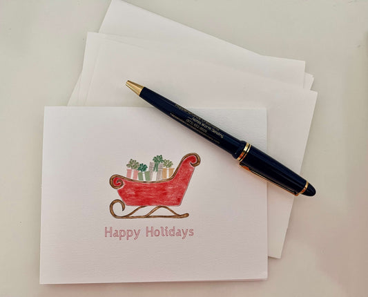Holiday note card with red sleigh illustration, 'Happy Holidays' text, and pen on white envelope