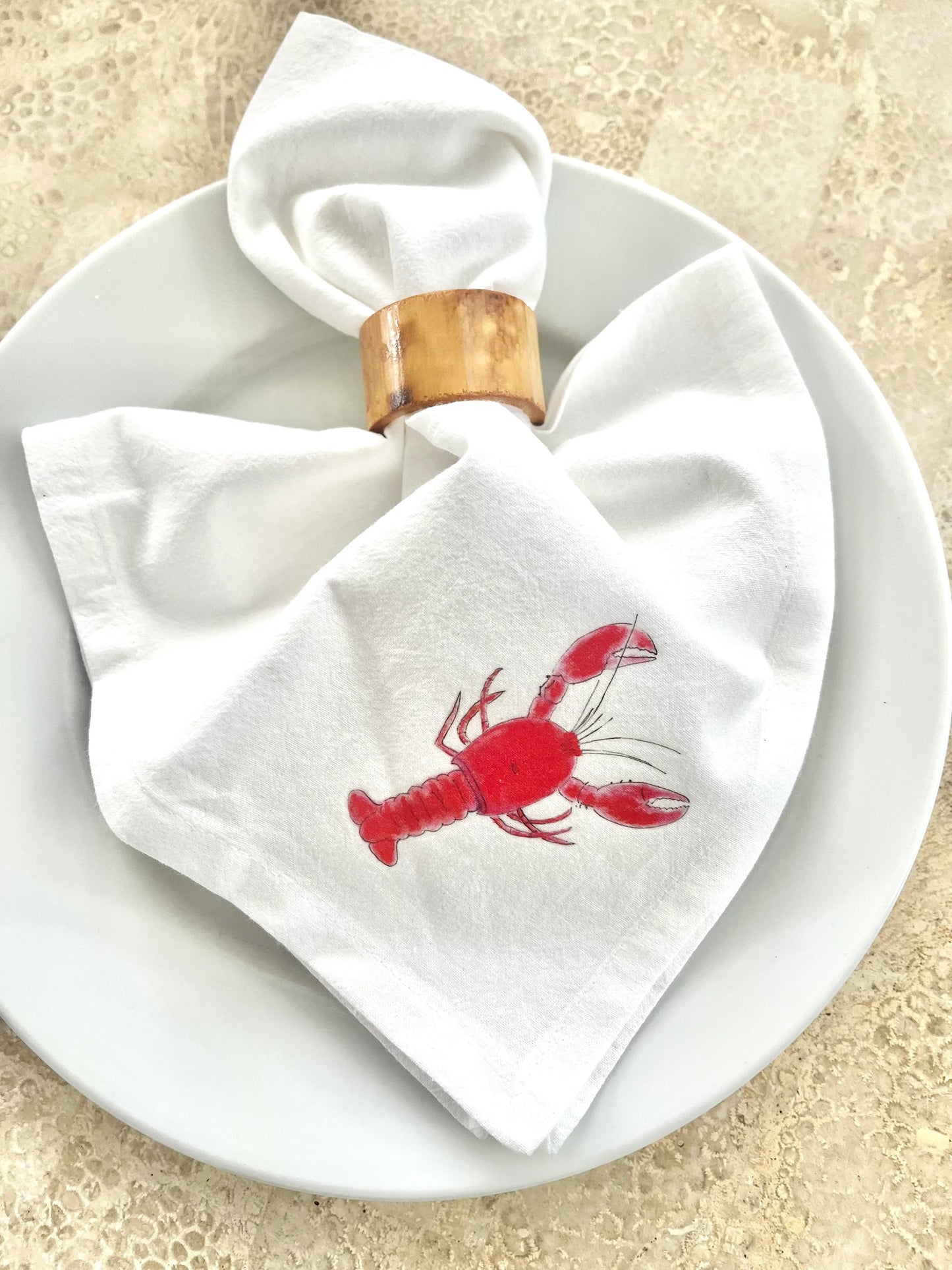 White cotton napkin with red lobster print, folded on white plate with wooden napkin ring, for coastal table settings.