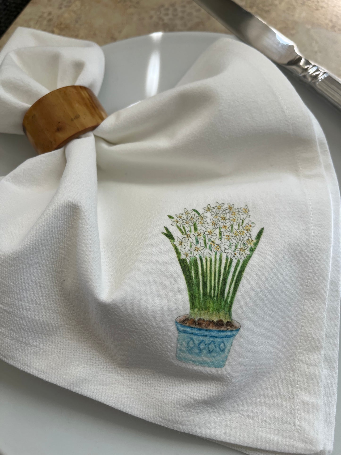 White cotton napkin with potted flower illustration, placed on a plate with a wooden napkin ring