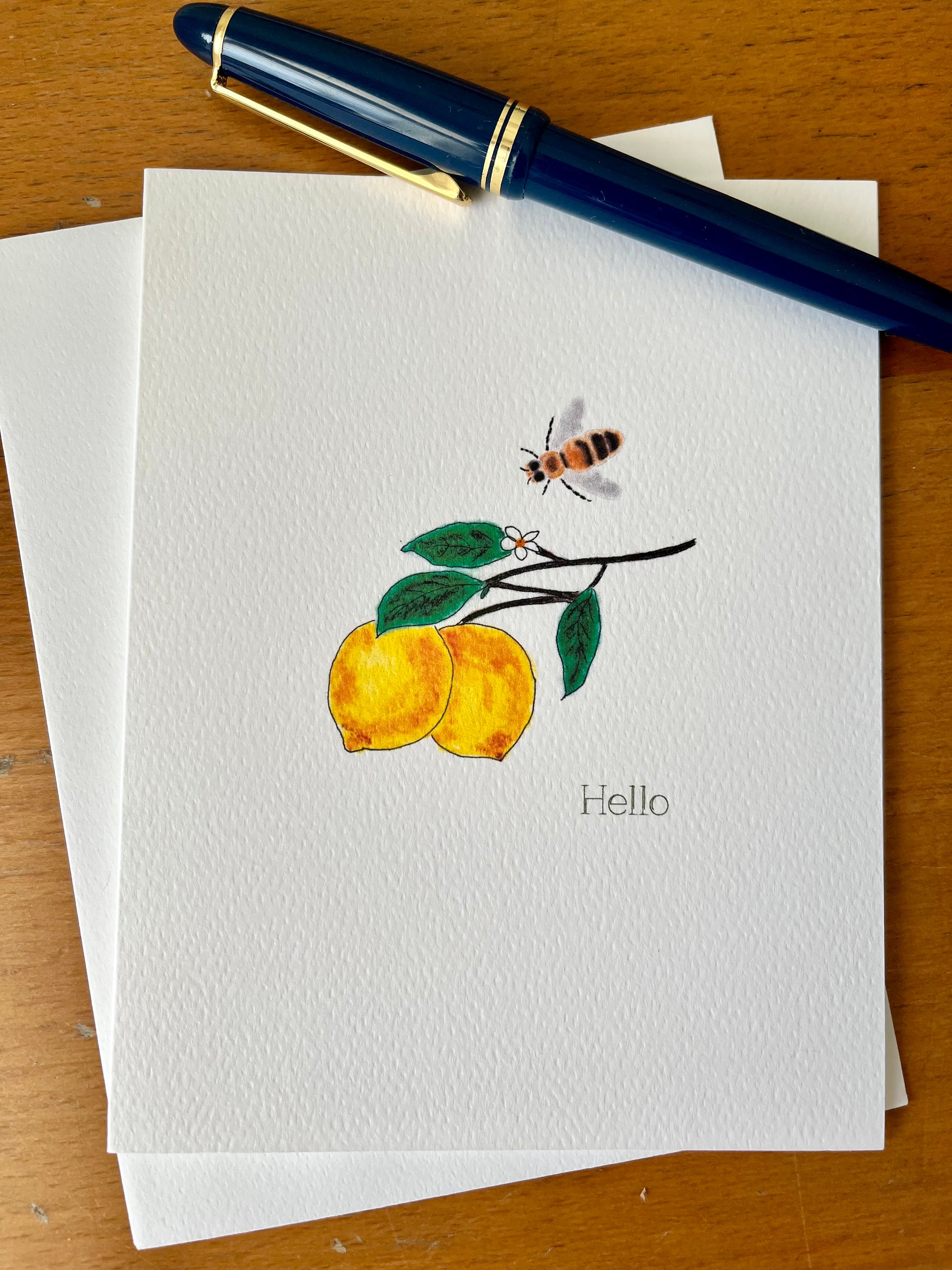 Illustrated note card with lemons, bee, and 'Hello' text on textured paper, with blue pen
