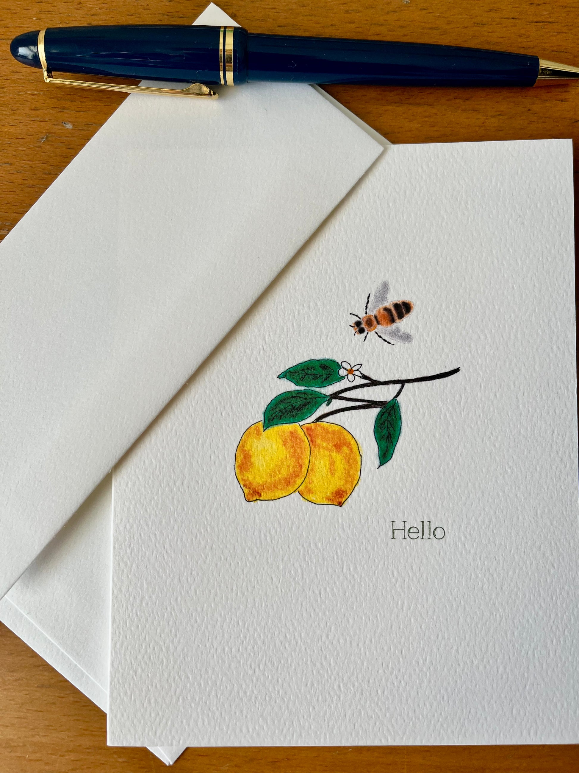 Illustrated lemon and bee note card with 'Hello' text, envelope, and blue pen on wood table.