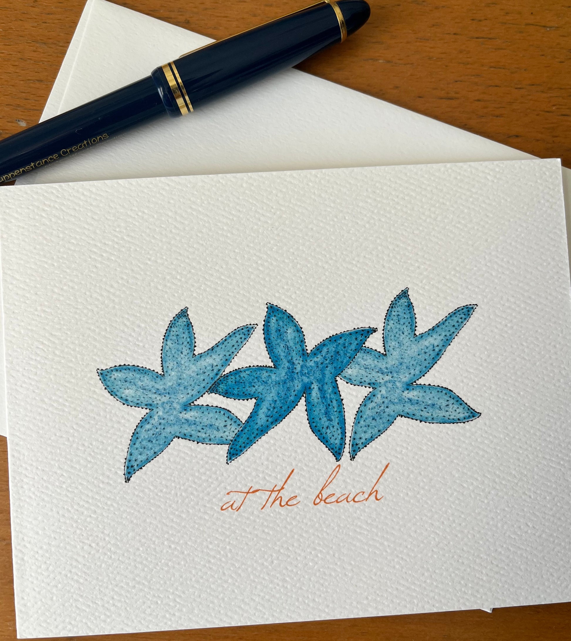 Note card with blue watercolor starfish illustration and 'at the beach' text, pen on table