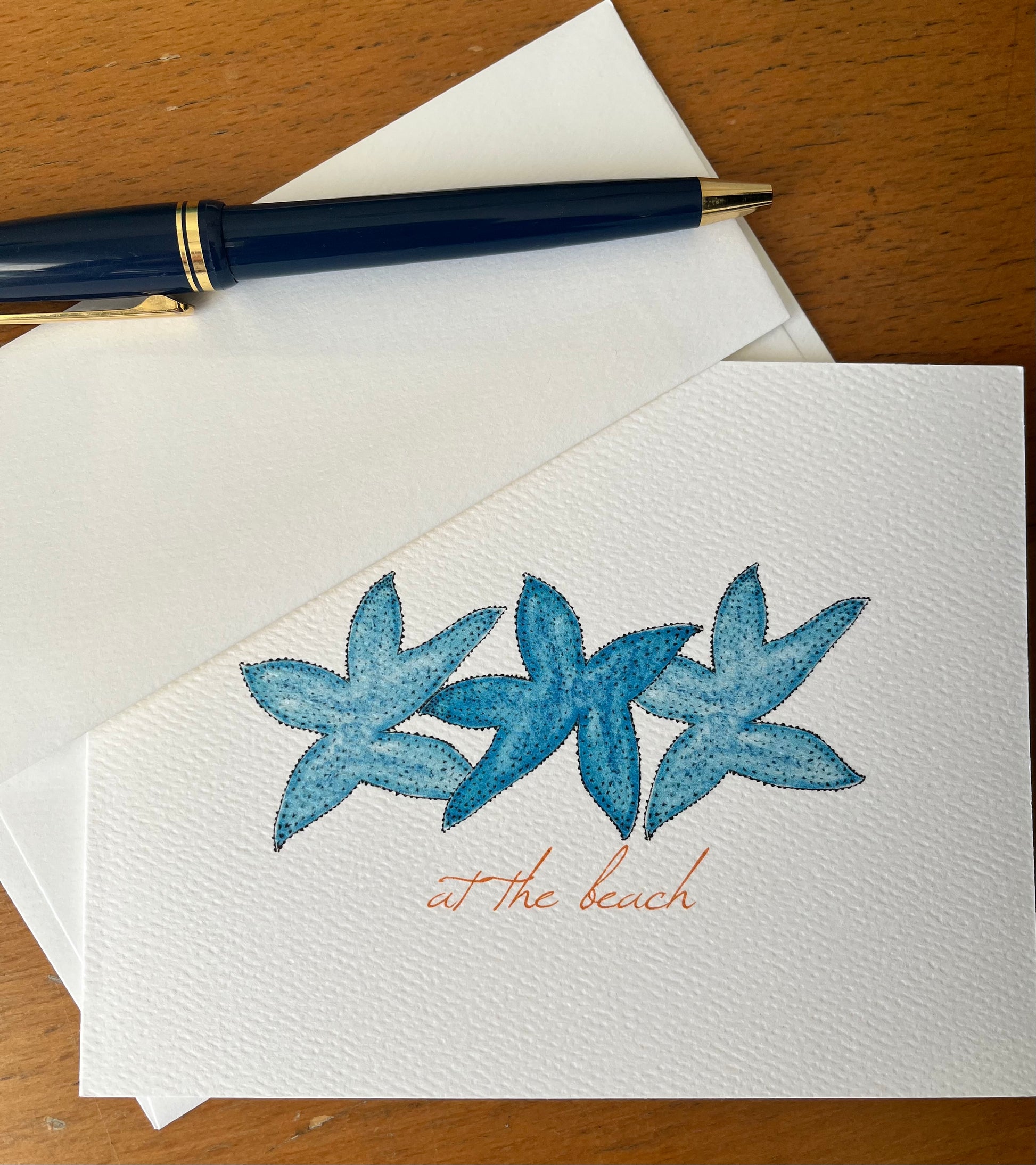 Illustrated starfish note card with 'at the beach' text, envelope, and blue pen on wood