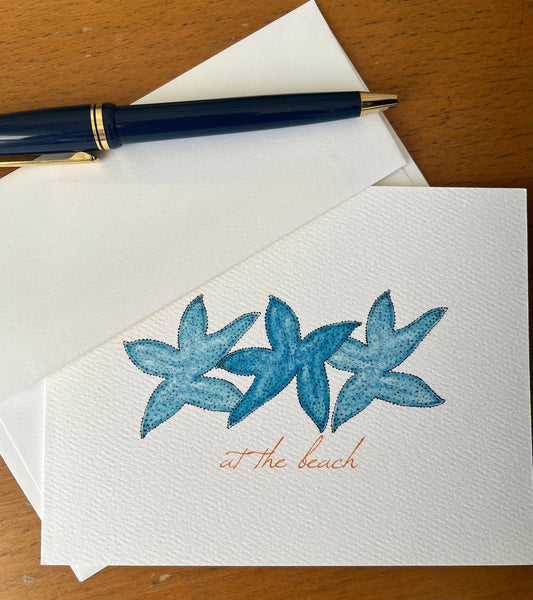 Illustrated starfish note card with 'at the beach' text, envelope, and blue pen on wood