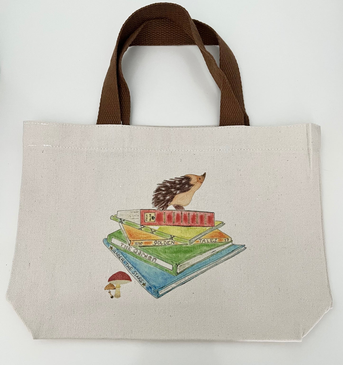 Canvas tote bag with brown handles, featuring a hedgehog illustration atop stacked books