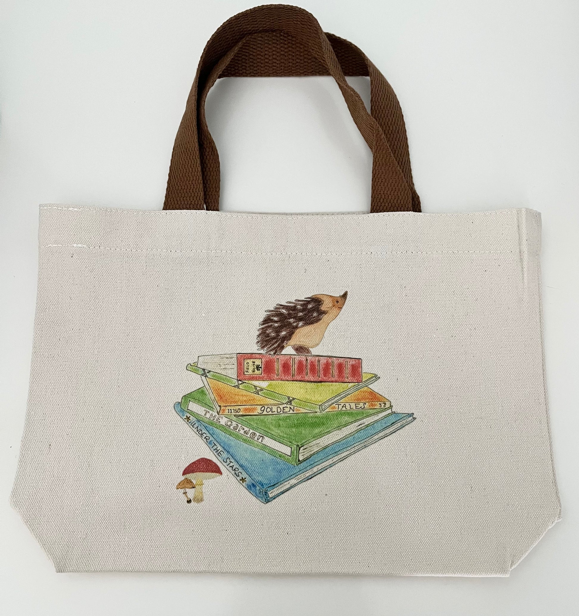 Canvas tote bag with brown handles, featuring a hedgehog illustration atop stacked books