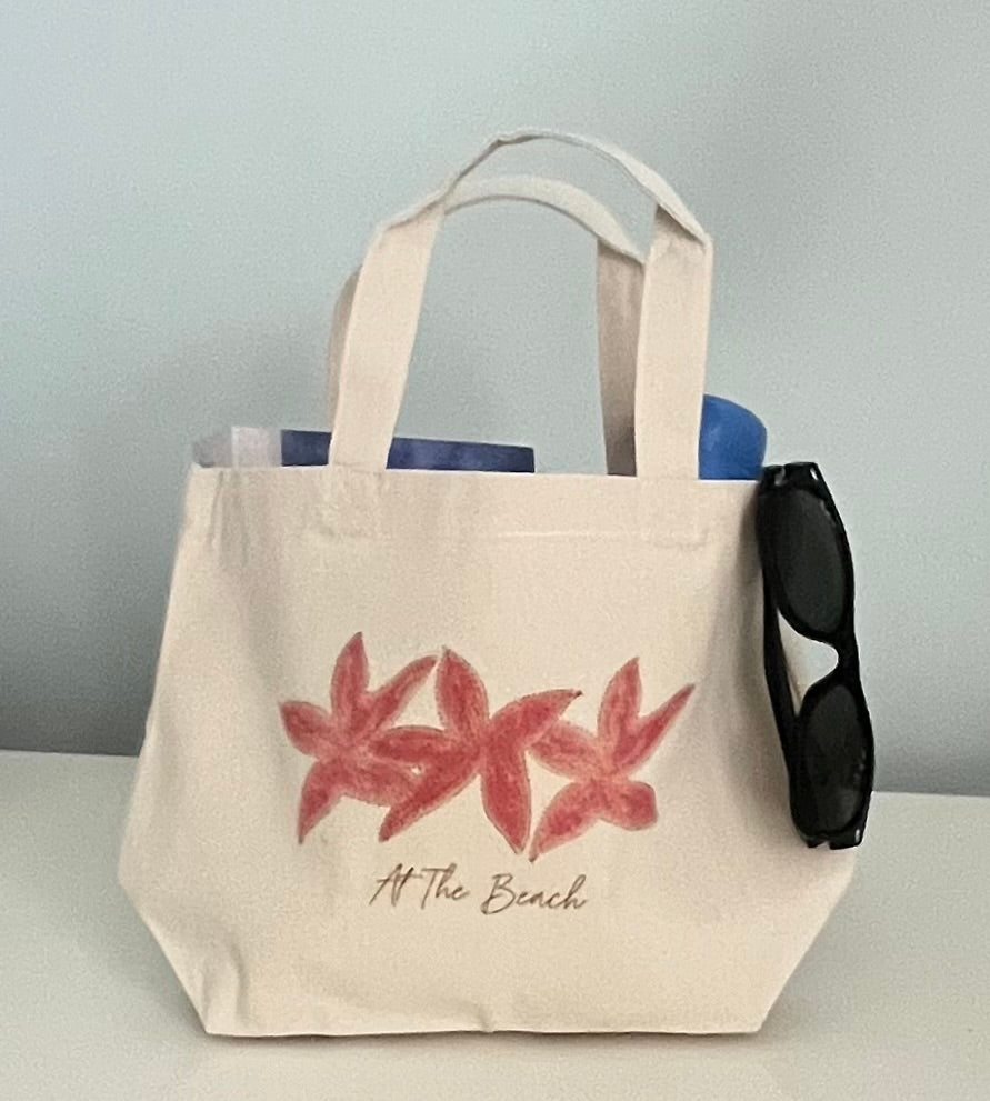 Beige tote bag with floral design and 'At The Beach' text, accompanied by sunglasses on a light gray background.