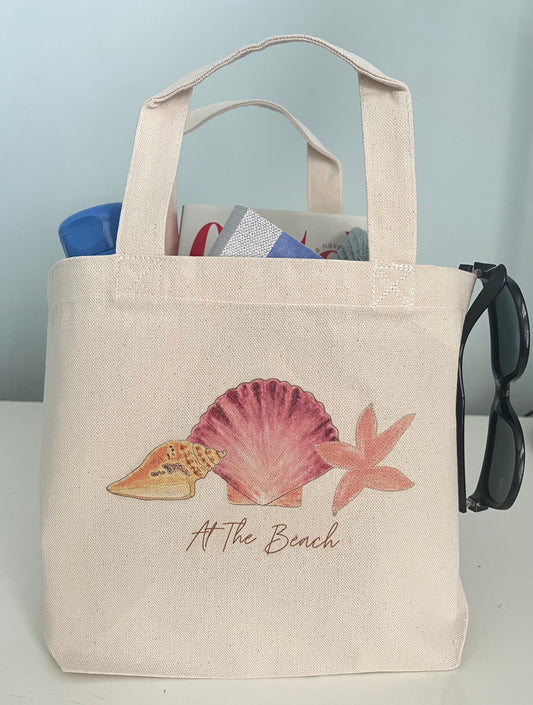 Beach tote bag with seashell, starfish, and 'At The Beach' text, perfect for seaside outings and nature-inspired gifts.