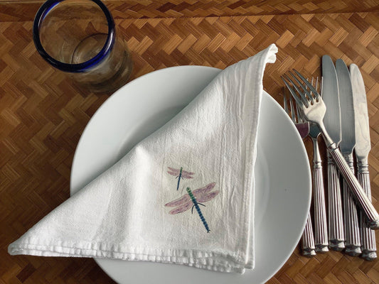 White cotton napkin with dragonfly print, set on plate with silverware and blue-rimmed glass, perfect for nature-inspired table settings.
