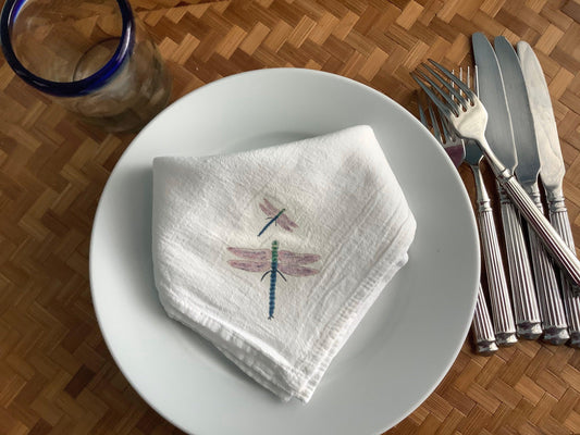 White cotton napkin with watercolor dragonfly design on dinner plate, silverware beside, nature-inspired table setting